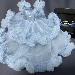 May include: A light blue tulle dress with ruffled trim and a beaded bodice. The dress is displayed on a gray surface with a black frame with the text "Sophie Toy Paradise" visible in the background.