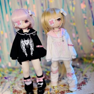K&ouml;nnte beinhalten: Zwei Puppen mit rosa und wei&szlig;en Outfits und Augenklappen. Die Puppe links tr&auml;gt ein schwarzes Hoodie mit wei&szlig;en Fl&uuml;geln und einer Kette. Die Puppe rechts tr&auml;gt ein wei&szlig;es Hoodie mit rosa Fl&uuml;geln und einer Kette.