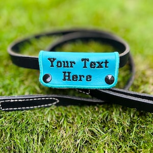 May include: A black leather dog leash with a turquoise handle featuring the embroidered text "Your Text Here". The handle is secured with black rivets. The leash is lying on green grass.