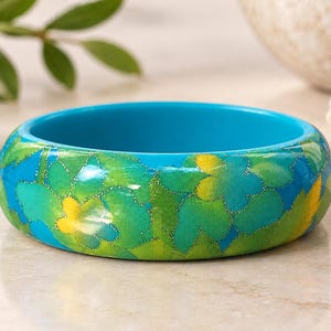 Puede incluir: Una pulsera tipo brazalete gruesa y redonda con un vibrante estampado floral en tonos azul brillante, verde lima y amarillo. La superficie exterior tiene un acabado brillante con sutiles detalles de purpurina que delinean las flores, mientras que el interior es de un color azul turquesa mate, liso y sólido.