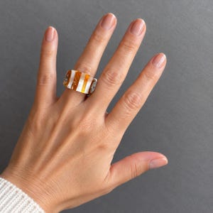 Può includere: Un anello a fascia larga caratterizzato da un motivo ripetitivo di strisce verticali color ambra e bianco opaco. L'anello presenta una finitura liscia e lucida e una forma rettangolare leggermente curva per adattarsi al dito.