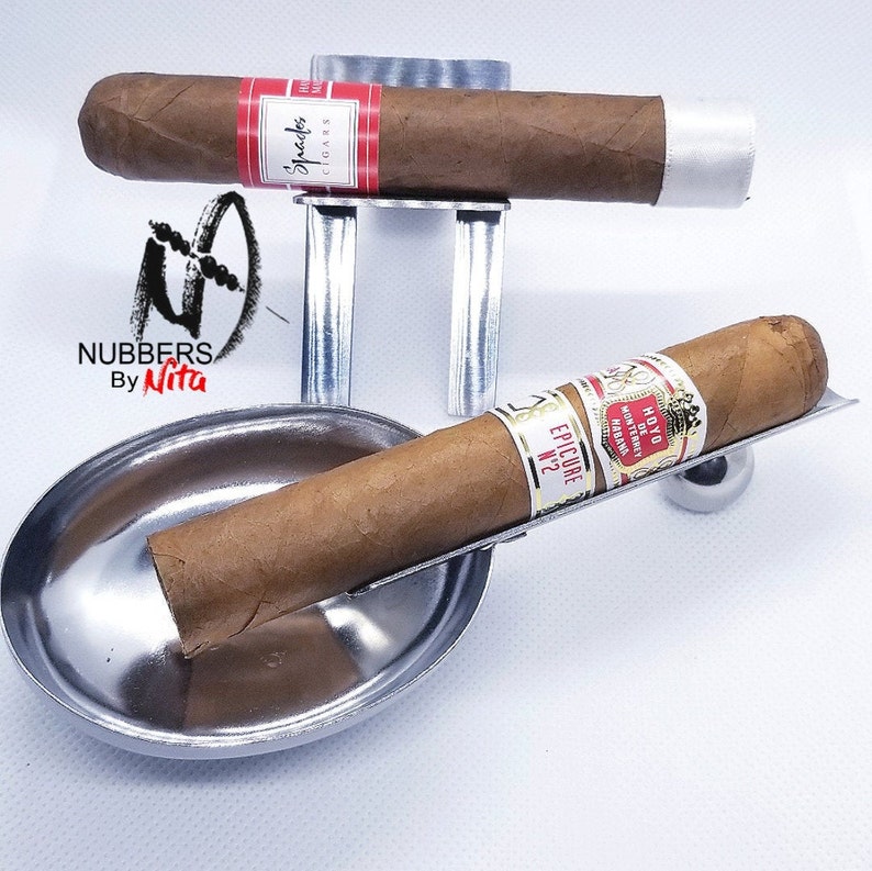 May include: Two silver cigar holders, one with a stand and one with a round ashtray. The cigar holders are designed to hold a cigar in place. The cigars are brown and have labels with text. The stand is made of metal and has a silver finish. The ashtray is also made of metal and has a silver finish.