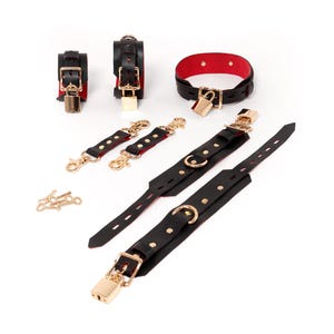Op de afbeelding: Een set zwarte lederen accessoires met rode voering en goudkleurige hardware, inclusief twee polsbanden, een halsband, twee korte verbindingsriemen, twee lange verstelbare riemen en een kleine verzameling goudkleurige sleutels voor de hangsloten.