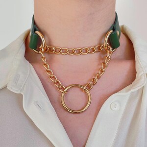 May include: A green leather choker necklace with gold-colored chain and ring accents. The necklace features a thick chain that connects to a large circular ring. The choker is worn on a person wearing a white button-down shirt.