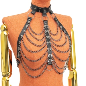 May include: Black faux leather body harness with silver-tone chain accents. The harness features a choker-style collar, shoulder straps, and cascading chains across the chest. The design includes metal rings and rivets for added detail. The harness is displayed on a mannequin.