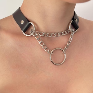 May include: Black leather choker necklace with silver-colored chain and ring accents. The choker features a wide band with a metal clasp and two large silver rings. A silver chain hangs from the rings, connecting to a larger ring in the center.