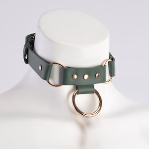 May include: A dark green leather choker with gold-toned metal rings and studs. The choker has a single large ring hanging from the bottom.