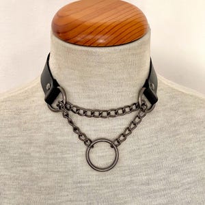 Op de afbeelding: Een zwarte leren choker met twee zilveren kettingen en een grote zilveren ring. De choker is op een mannequin.