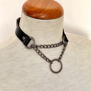 Könnte beinhalten: Ein schwarzes Lederhalsband mit zwei silbernen Ketten und einem großen silbernen Ring. Das Halsband befindet sich auf einer Puppe.