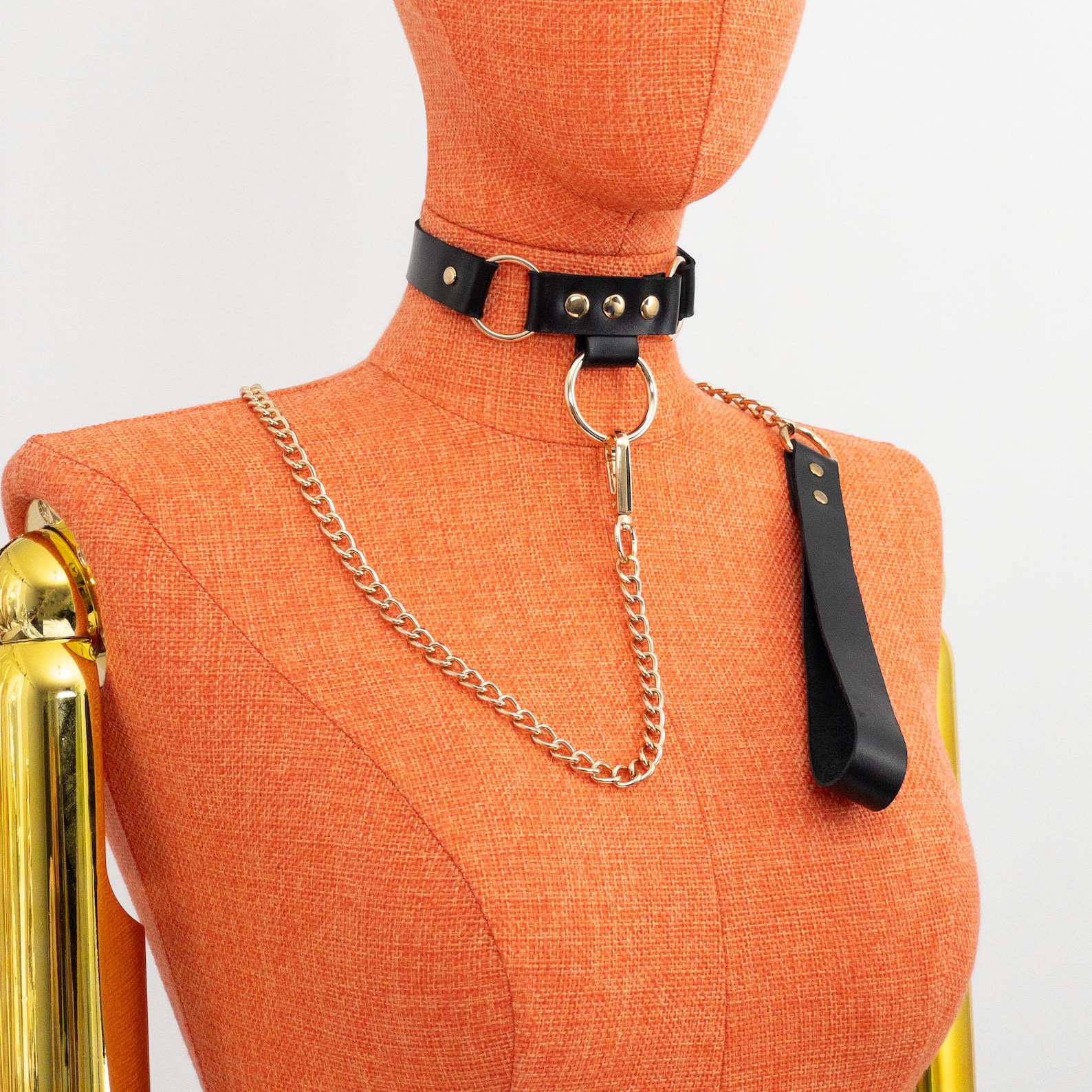 Black leather bdsm collar with gold chain leash on orange mannequin, featuring O-ring detail and sleek bondage style.