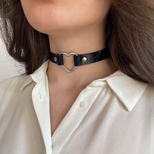 May include: Black choker necklace with a silver heart pendant. The choker is made of black leather and has silver-toned metal hardware. The heart pendant is a hollow, open design. The necklace is worn around the neck.