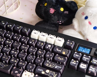 May include: Black mechanical keyboard with cat-themed keycaps. The keycaps feature cat faces and other cat-related designs. The keyboard has a small screen and a volume control knob. Two plush cat toys are in the background.