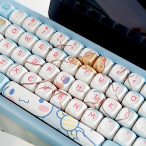 May include: A light blue mechanical keyboard with custom keycaps. The keycaps are white with various cartoon character designs and pastel colors. The keyboard has a light blue frame.