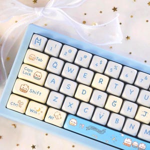 May include: A light blue mechanical keyboard with white and cream-coloured keycaps. The keycaps have light blue lettering and cute character illustrations. A pair of decorative hair clips with cartoon characters are also in the image.