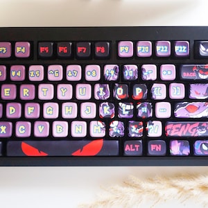 May include: A black mechanical keyboard with custom keycaps. The keycaps are a mix of pink, purple, and black, featuring various designs and text. The keyboard has a volume control knob and a small display. The overall theme is a custom gaming keyboard.