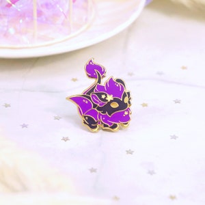 May include: A purple and black enamel pin featuring a stylized creature with gold accents. The pin has a gold border and star details. The background is a soft, white fabric with scattered star shapes.