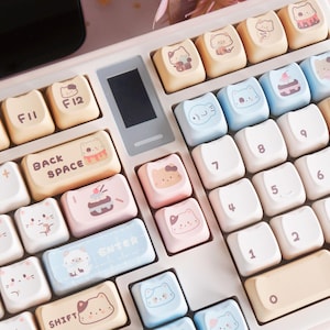 以下が含まれることがあります： 白とピンクのメカニカルキーボード。キーキャップは、パステルカラーのかわいい漫画の猫とデザートのデザインが特徴です。キーボードにはテンキーとファンクションキーがあり、「BACK SPACE」と「SHIFT」の文字が見えます。