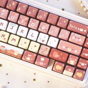 May include: A white and pink mechanical keyboard with custom keycaps. The keycaps feature bear and paw designs in shades of pink and white. The keyboard has a decorative border with bear illustrations. The keyboard is on a white surface.
