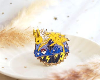 May include: A yellow and white Jolteon enamel pin with a blue and gold background. The pin features a lightning bolt design and a Pokeball. The pin is round with a gold border and is set on a white plate.