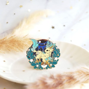 Puede incluir: Un pin de esmalte dorado con una criatura mítica de color amarillo claro con detalles azules y verdes. La criatura está rodeada de estrellas y una luna creciente. El pin está sobre un fondo blanco.