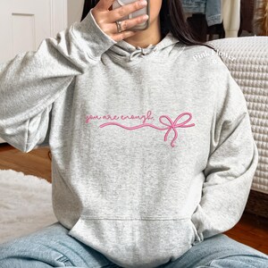 May include: A light gray hoodie with a front pocket and a hood. The hoodie features the embroidered phrase "you are enough" in pink script, accompanied by a pink bow design. The model is wearing blue jeans.