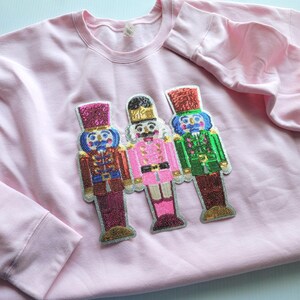 May include: A light pink sweatshirt featuring three beaded nutcracker figures. The nutcrackers are in red, pink, and green uniforms with matching hats. The sweatshirt has long sleeves and a crew neck.