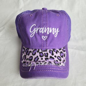 May include: Purple baseball cap with the word "Granny" embroidered in white script, a small heart, and a leopard print brim. The cap has a mesh back and a curved brim.