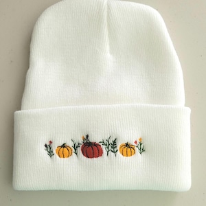 Op de afbeelding: Witte gebreide beanie met een geborduurd ontwerp van pompoenen en bladeren in oranje, geel en groen.