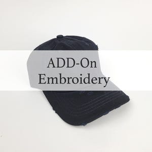 May include: A navy blue baseball cap with distressed detailing. The cap features the text "ADD-On Embroidery" in a white overlay. The hat has a curved brim and a button on top. The background is plain white.