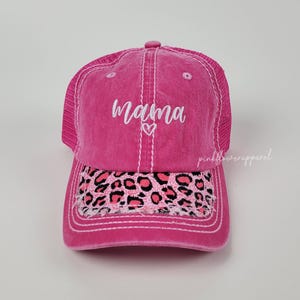 May include: A pink trucker hat with the word "mama" embroidered in white script above a small heart. The brim features a leopard print design in pink and black. The hat has a mesh back and a curved brim.