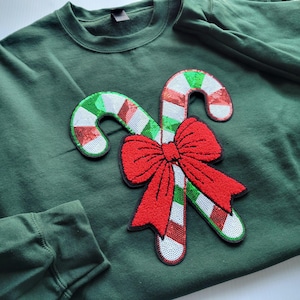 May include: A dark green sweatshirt featuring a festive design. Two candy canes with red, white, and green sequins are paired with a large red sequined bow. The sweatshirt is a comfortable, long-sleeved garment.