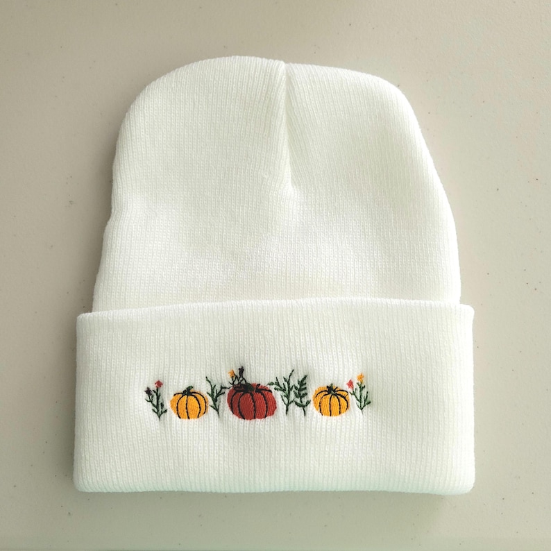 Op de afbeelding: Witte gebreide beanie met een geborduurd ontwerp van pompoenen en bladeren in oranje, geel en groen.