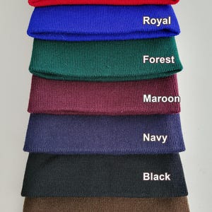 Op de afbeelding: Een stapel van acht gebreide mutsen in verschillende kleuren: rood, koninklijk blauw, bosgroen, donkerrood, marineblauw, zwart en bruin. De kleuren zijn op elke muts gelabeld.