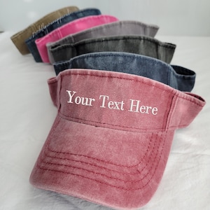 May include: A row of visors in various colors, including dusty rose, gray, and denim blue. The front visor is dusty rose with the text "Your Text Here" embroidered in white. The visors are made of a soft, textured material.