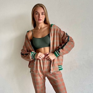 May include: A woman is wearing a pink and green checkered tracksuit with a white zipper. She is wearing a green tank top underneath the jacket. She is wearing white sneakers with green accents.