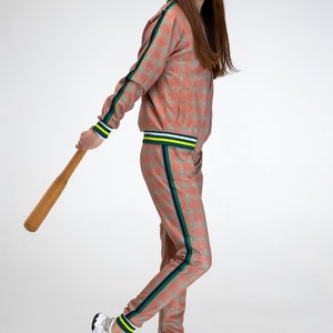 May include: A person wearing a pink and green checked tracksuit with a white and yellow stripe down the side of the trousers. They are holding a wooden baseball bat and wearing white trainers.