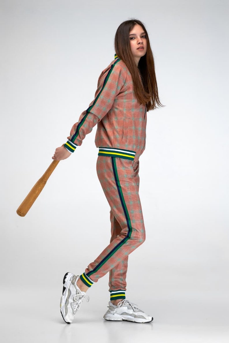 May include: A person wearing a pink and green checked tracksuit with a white and yellow stripe down the side of the trousers. They are holding a wooden baseball bat and wearing white trainers.