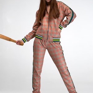 May include: A woman wearing a pink and green checked tracksuit with yellow and green stripes on the cuffs. She is holding a wooden baseball bat.