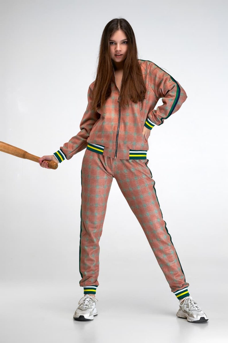 May include: A woman wearing a pink and green checked tracksuit with yellow and green stripes on the cuffs. She is holding a wooden baseball bat.