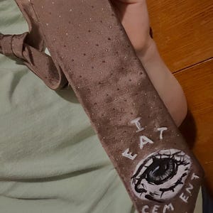 Può includere: Una cravatta marrone con pois bianchi e un occhio dipinto con le parole "I EAT CEMENT" su di essa.