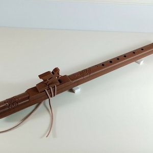 Puede incluir: Una flauta de madera marrón con un diseño tallado y seis agujeros para los dedos. La flauta está atada con un cordón de cuero marrón y descansa sobre dos bloques blancos.