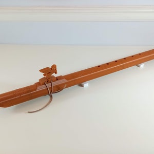 May include: A brown wooden flute with a bird carving on top. The flute has six finger holes and is resting on two white blocks.