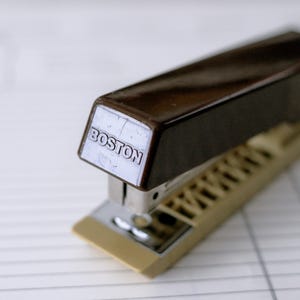 May include: A vintage brown and tan stapler with the word "BOSTON" printed on the top. The stapler has a metal base and a dark brown top. The stapler is on a white surface.