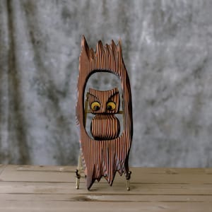 May include: A wooden owl sculpture with a cutout design. The owl is brown with yellow eyes and is set within a frame that resembles a tree trunk. The sculpture is supported by small, gold-colored legs.