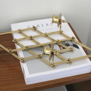 May include: A gold-toned metal accordion-style wall rack featuring a lattice design with small circular knobs at each intersection and two cylindrical spool-like attachments extending outward from the frame, resting on a white magazine.