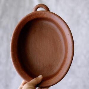 May include: A round, terracotta-colored clay dish with a handle. The dish has a shallow, flat interior and a raised rim. The handle is a small, arched loop. The dish is held by a hand against a neutral background.