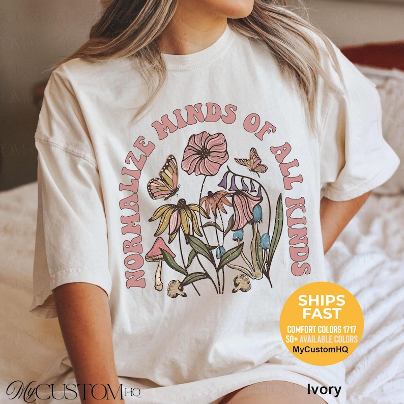 May include: An ivory t-shirt featuring a graphic print with the text NORMALIZE MINDS OF ALL KINDS in pink letters. The design includes illustrations of various flowers, mushrooms, and butterflies in shades of pink, yellow, blue, and green.