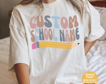 May include: An ivory short-sleeve t-shirt featuring the text CUSTOM SCHOOL NAME in a wavy, multi-colored font of pink, blue, and tan. Below the text is a graphic of a yellow pencil with a pink eraser and a grey ferrule, centered on the chest.