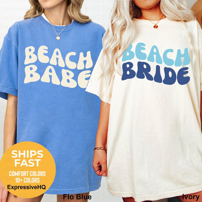 Puede incluir: Una camiseta azul con el texto BEACH BABE en blanco y una camiseta color marfil con el texto BEACH BRIDE en azul claro y oscuro. Ambas camisetas presentan cuello redondo y mangas cortas, mostrando un estilo casual con tipograf&iacute;a ondulada.