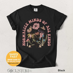 May include: A black short-sleeve t-shirt featuring a graphic print of flowers, butterflies, and mushrooms arranged in an arch. The text NORMALIZE MINDS OF ALL KINDS is printed in a light pink arched font around the central botanical illustration.